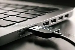 A close-up black and white photo of a laptop's side view with a USB cable plugged in. The laptop keyboard is partially visible, connecting the USB connection